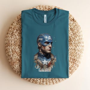 May include: Teal t-shirt featuring a Captain America graphic. The illustration depicts Captain America in his iconic blue and white costume, with a stern expression. The shirt is folded on a woven, circular surface.