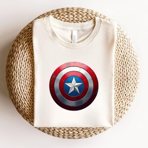May include: A cream-coloured crew neck t-shirt featuring a centred graphic of a circular shield with a distressed red, white, and blue finish. The shield design includes a central white star on a blue background, surrounded by concentric rings.