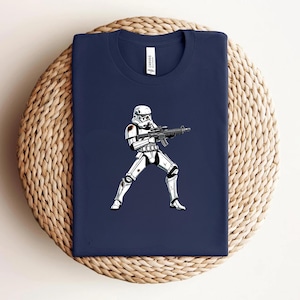May include: Navy blue t-shirt featuring a white and black illustration of a Stormtrooper holding a blaster. The graphic is centered on the shirt, which is folded on a woven surface. The shirt has a crew neck.