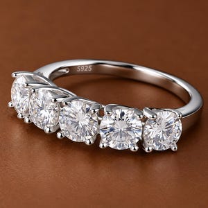 Puede incluir: Un anillo de tono plateado con cinco piedras redondas, transparentes y facetadas dispuestas en fila. La banda es de metal pulido y tiene el grabado S925 claramente visible en la superficie interior, indicando la composición del material.