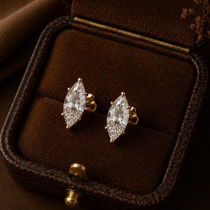May include: A pair of stud earrings featuring marquise-cut clear stones set in yellow gold. Each earring has a pointed, elongated shape with multiple prongs securing the center stone. The earrings are displayed inside a dark brown velvet box.