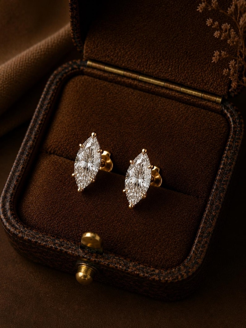 May include: A pair of stud earrings featuring marquise-cut clear stones set in yellow gold. Each earring has a pointed, elongated shape with multiple prongs securing the center stone. The earrings are displayed inside a dark brown velvet box.