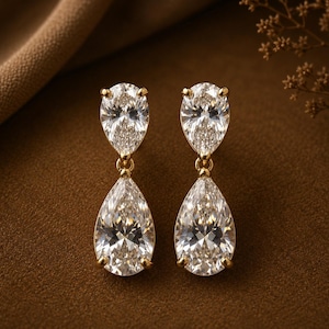 Peut inclure: Une paire de boucles d'oreilles pendantes en or avec deux pierres transparentes en forme de poire chacune. La pierre supérieure est plus petite et reliée à une pierre plus grande en forme de poire par un petit anneau en or.