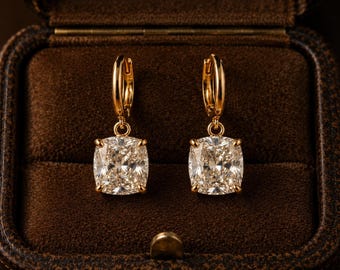 May include: A pair of gold hoop earrings featuring cushion-cut clear gemstones suspended from each hoop. The earrings are displayed symmetrically, showcasing the polished gold metal finish and the faceted, brilliant-cut appearance of the stones.