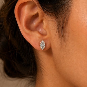 May include: A marquise-cut clear gemstone stud earring set in a gold-toned metal prong setting. The earring is positioned in the earlobe, featuring a faceted, elongated oval shape that tapers to points at both ends, reflecting light clearly.