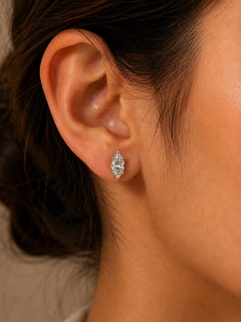 May include: A marquise-cut clear gemstone stud earring set in a gold-toned metal prong setting. The earring is positioned in the earlobe, featuring a faceted, elongated oval shape that tapers to points at both ends, reflecting light clearly.