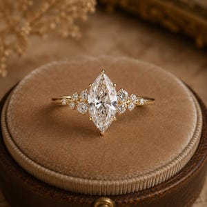 May include: A gold ring featuring a large marquise-cut centre stone flanked by a cluster of smaller round-cut stones on each side. The band is thin and polished, holding the central marquise diamond securely in a prong setting with accent stones.