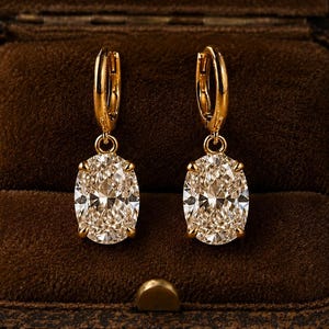 May include: A pair of gold hoop earrings featuring oval-cut clear stones suspended from the base. Each earring has a polished gold metal hoop mechanism at the top, connecting to a prong-set oval stone that reflects light with a faceted surface.