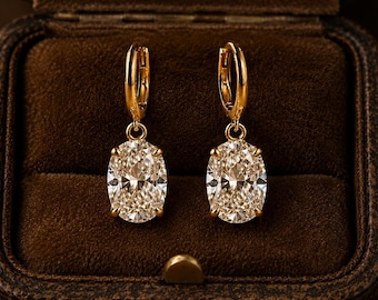 May include: A pair of gold hoop earrings featuring oval-cut clear stones suspended from the base. Each earring has a polished gold metal hoop mechanism at the top, connecting to a prong-set oval stone that reflects light with a faceted surface.