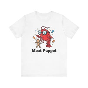 May include: White t-shirt featuring a cartoon red lobster wearing glasses and a small wooden puppet. The text "Meat Puppet" is printed below the graphic. The lobster is surrounded by blue and orange lightning bolts.