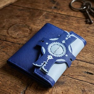 Può includere: Un portafoglio blu con un accento azzurro e una chiusura decorativa. La chiusura presenta un design circolare con un motivo a ingranaggi e testo. Il portafoglio è su una superficie di legno.