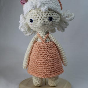 May include: A handmade crochet doll with a white hat adorned with yellow and pink flowers. The doll has blue eyes and wears a peach-colored dress. The doll is standing on a wooden base.