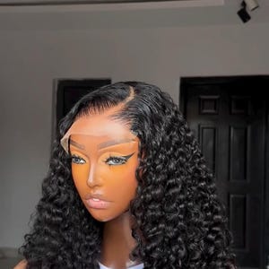 May include: A black curly wig with a lace front. The wig is styled with a side parting and has a natural-looking hairline. The person is wearing a white vest top with the word "HOT" printed on it.