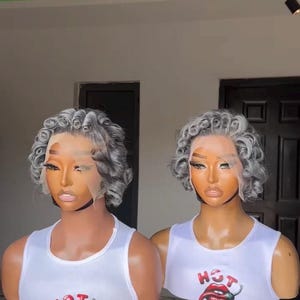 May include: Two mannequin heads with gray curly wigs and lace fronts are displayed. They wear white tank tops with a red and black graphic that says "HOT" with a chili pepper and lips. The mannequins are mounted on tripods.