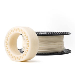 Puede incluir: Primer plano de dos bobinas de filamento de impresión 3D. Una es un objeto en forma de rosquilla de color crema con una serie de recortes triangulares. La otra es una bobina negra que contiene un rollo de filamento crema. El fondo es blanco.