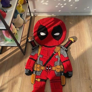 Puede incluir: Una alfombra roja de Deadpool con formas de ojos negros y acentos de ojos blancos. La alfombra presenta un diseño detallado del disfraz del personaje, que incluye un cinturón de herramientas, espadas y acentos grises y amarillos. La alfombra está colocada sobre un suelo de madera.