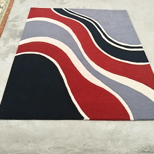 May include: A rectangular area rug featuring a bold, abstract pattern of flowing, wavy bands in deep navy blue, vibrant red, soft light gray, and cream. The design consists of thick, curved stripes that sweep across the surface in a rhythmic, undulating motion, creating a dynamic visual effect with high-contrast color blocking throughout the entire piece.