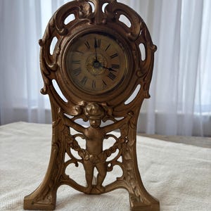 Puede incluir: Reloj de sobremesa antiguo de color dorado con números romanos en la esfera. El reloj tiene un diseño ornamentado con una figura de querubín en la base y adornos decorativos. Una pieza vintage.