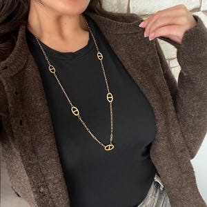 Peut inclure: Un long collier doré avec des maillons ovales est présenté. Le collier est porté sur un haut noir et associé à un cardigan marron. Le bijou est un accessoire simple mais élégant.