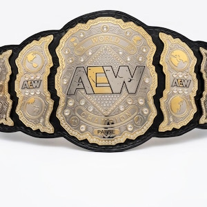 May include: A professional wrestling championship belt with a black leather strap and gold and silver metal plates. The center plate features the letters "AEW" and the words "WORLD CHAMPION". The side plates have the letters "AEW" and a globe design.
