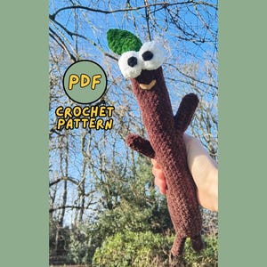 Op de afbeelding: Een bruin gehaakt stokfiguur met witte ogen, een groen blad en een lachend gezicht. De figuur heeft armen en benen. De afbeelding bevat de tekst "PDF CROCHET PATTERN" in een cirkel.