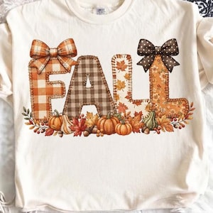 Puede incluir: Una camiseta de color crema con la palabra FALL en letras grandes estilo costura. Cada letra tiene un patrón único: cuadros naranjas, vichy marrón, hojas de otoño y estampado floral. El diseño incluye lazos y calabazas.