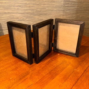 May include: A triptych photo frame featuring three connected rectangular frames made of dark brown wood. The frames are hinged together, allowing them to stand in a zigzag arrangement, each containing a plain beige insert behind clear glass.