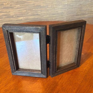 May include: A double hinged picture frame constructed from dark brown wood with a visible grain texture. The two rectangular frames are connected by two black metal hinges, allowing the unit to stand upright in a V-shaped configuration.