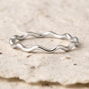 Può includere: Un sottile anello in argento lucido caratterizzato da un design ondulato o serpentino continuo e ripetitivo. La fascia in metallo ha una superficie liscia e riflettente che cattura la luce, creando una serie di curve che formano l'anello.
