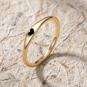 May include: A gold ring featuring a black heart-shaped detail. The ring has a sleek, polished appearance and a slender band. The heart is inlaid into the ring's surface. The ring is displayed against a textured, neutral-toned backdrop.