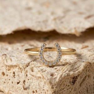 Puede incluir: Un anillo de oro con un diseño de herradura incrustado con pequeños diamantes brillantes. El anillo se presenta sobre un fondo texturizado de tonos neutros, resaltando los detalles de la joya. La herradura es un símbolo de buena suerte.