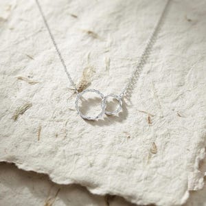 Può includere: Una collana in argento con due pendenti circolari intrecciati con una superficie ritorta e testurizzata. La catena sottile si collega al cerchio più grande, che è unito a un cerchio più piccolo, creando un design continuo e collegato.