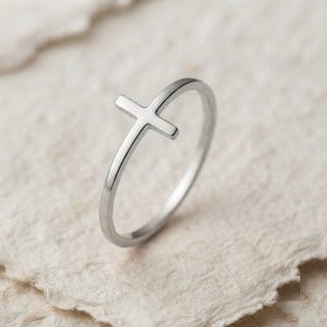 May include: A thin, polished silver-tone metal ring featuring a small, upright cross design integrated into the band. The ring has a smooth, reflective surface and a simple, continuous circular shape, showcasing a clean and minimalist aesthetic.