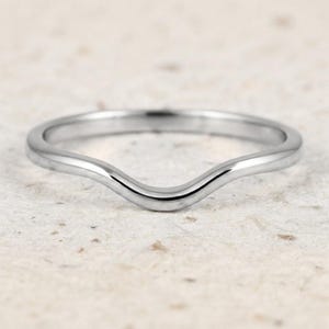 May include: A polished silver-toned metal ring featuring a smooth, continuous band with a distinct V-shaped curve or chevron contour at the centre. The ring has a reflective, metallic finish and a simple, unadorned design throughout the entire band.