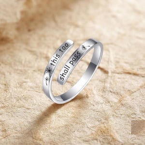 Puede incluir: Un anillo abierto de tono plateado con el texto grabado "this too shall pass" y pequeñas ilustraciones de semillas de diente de león. La banda de metal tiene un acabado liso y pulido, curvándose para formar un diseño ajustable y abierto.