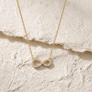 Puede incluir: Un collar dorado con un colgante con el símbolo del infinito. El símbolo del infinito está engastado con pequeñas piedras preciosas transparentes. La cadena es delicada y también dorada. La joya se exhibe sobre una superficie texturizada de color blanquecino.