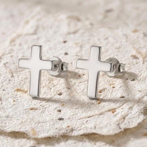 May include: A pair of silver cross stud earrings. The earrings are presented on a textured, off-white surface. Each earring features a simple, polished cross design.
