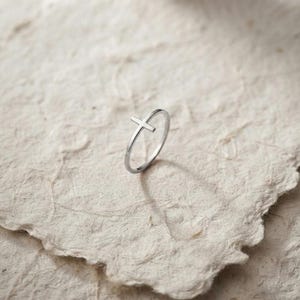 May include: A thin, polished silver-tone ring featuring a small, centred cross design. The band is smooth and circular, with the cross element positioned upright. The metal has a reflective, metallic finish that catches the light, creating a clean, simple look.