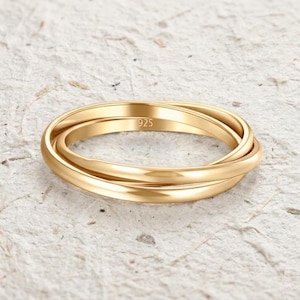May include: A gold-colored ring with three interlocking bands. The ring has a polished finish and the number "925" is engraved on one of the bands. The ring is set against a textured, off-white background.