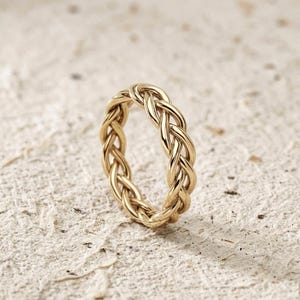 May include: A gold ring featuring a braided design. The ring's interwoven pattern creates a textured appearance. The ring is set against a neutral background, highlighting its intricate details. This piece is a jewelry item.