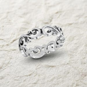 Puede incluir: Un anillo de tono plateado que presenta un intrincado diseño de filigrana calada con patrones de volutas que envuelven toda la circunferencia de la banda, creando una estructura metálica decorativa y continua con superficies pulidas.
