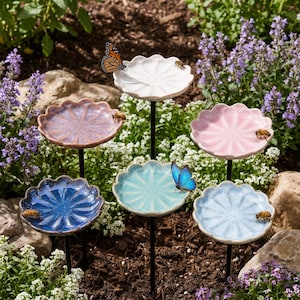 May include: A set of six ceramic flower-shaped garden stakes in various colours including white, pink, light blue, dark blue, teal, and purple. Each dish-shaped top is mounted on a thin black metal rod, with butterflies and bees resting on them.