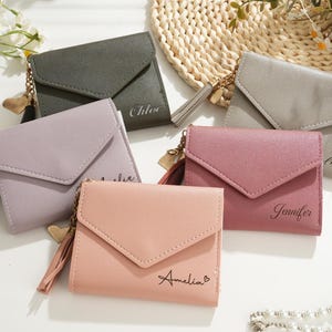 Puede incluir: Cinco carteras pequeñas y personalizadas en varios colores pastel: rosa empolvado, lavanda, verde oliva, gris y malva. Cada cartera tiene un cierre de solapa, una borla y un nombre impreso en la parte delantera. Un dije de corazón dorado cuelga de la cremallera.