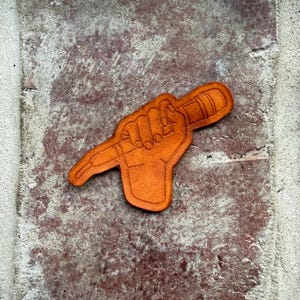 May include: An orange leather patch depicting a hand holding a microphone. The patch has a detailed outline and is a solid colour. The background is a textured, reddish-brown surface, similar to brick.
