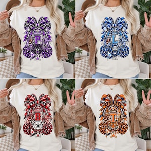 Può includere: Una t-shirt color crema con una grafica composta da un grande fiocco, una mascotte sportiva, pon pon e un megafono. Sono mostrate quattro varianti: viola con una pantera, blu con un leone, rosso con un bulldog e arancione con una tigre.