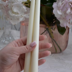 May include: Two ivory-colored taper candles. The candles are held in front of a blurred background of pink and white flowers in a vase. The candles are long and slender, with a smooth surface.