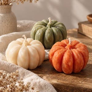 May include: Three pumpkin-shaped candles in cream, sage green, and burnt orange colors. Each candle features a ribbed texture and a single central wick. The candles are arranged on a wooden surface with a textured fabric cloth draped underneath.