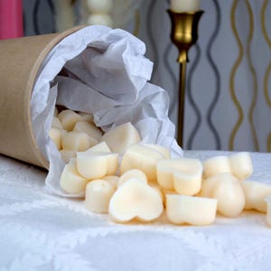 May include: A brown cardboard container tipped over, spilling heart-shaped wax melts onto a white tablecloth. The melts are a creamy off-white color. The container is lined with white tissue paper. The background includes a gold candlestick and patterned wallpaper.