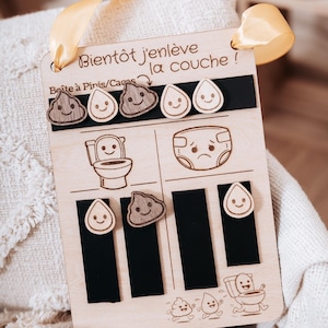 Peut inclure: Tableau d'apprentissage de la propreté en bois avec le texte "Bientôt j'enlève la couche !" et des illustrations de toilettes, de couches et de formes de caca et de goutte d'eau souriantes. Comprend un ruban jaune pour l'accrocher.