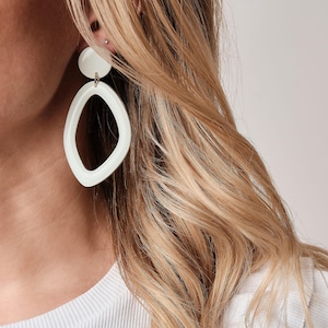 Könnte beinhalten: Weiße, geometrische Ohrringe mit einem kreisförmigen Oberteil und einem ovalen Unterteil. Die Ohrringe bestehen aus einem festen, undurchsichtigen Material und hängen an einem goldfarbenen Haken. Die Ohrringe werden vor blondem Haar und einem weißen Hemd gezeigt.